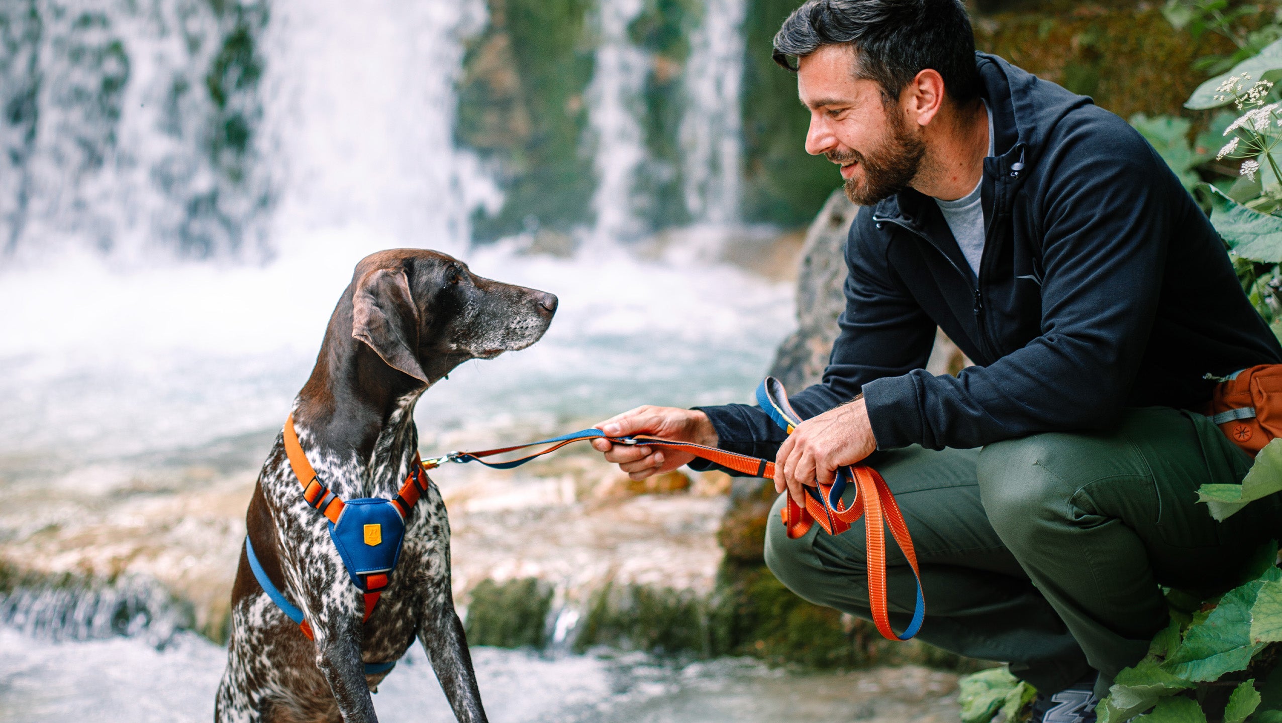 Man kneeling by a waterfall, smiling at a dog on a leash, surrounded by greenery.