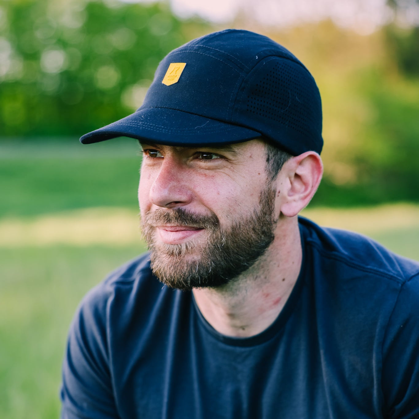 A bearded man wearing the Alpha 360 Tech Cap Black and a navy t-shirt sits on a grassy field outdoors, with trees and sunlight in the blurred background.