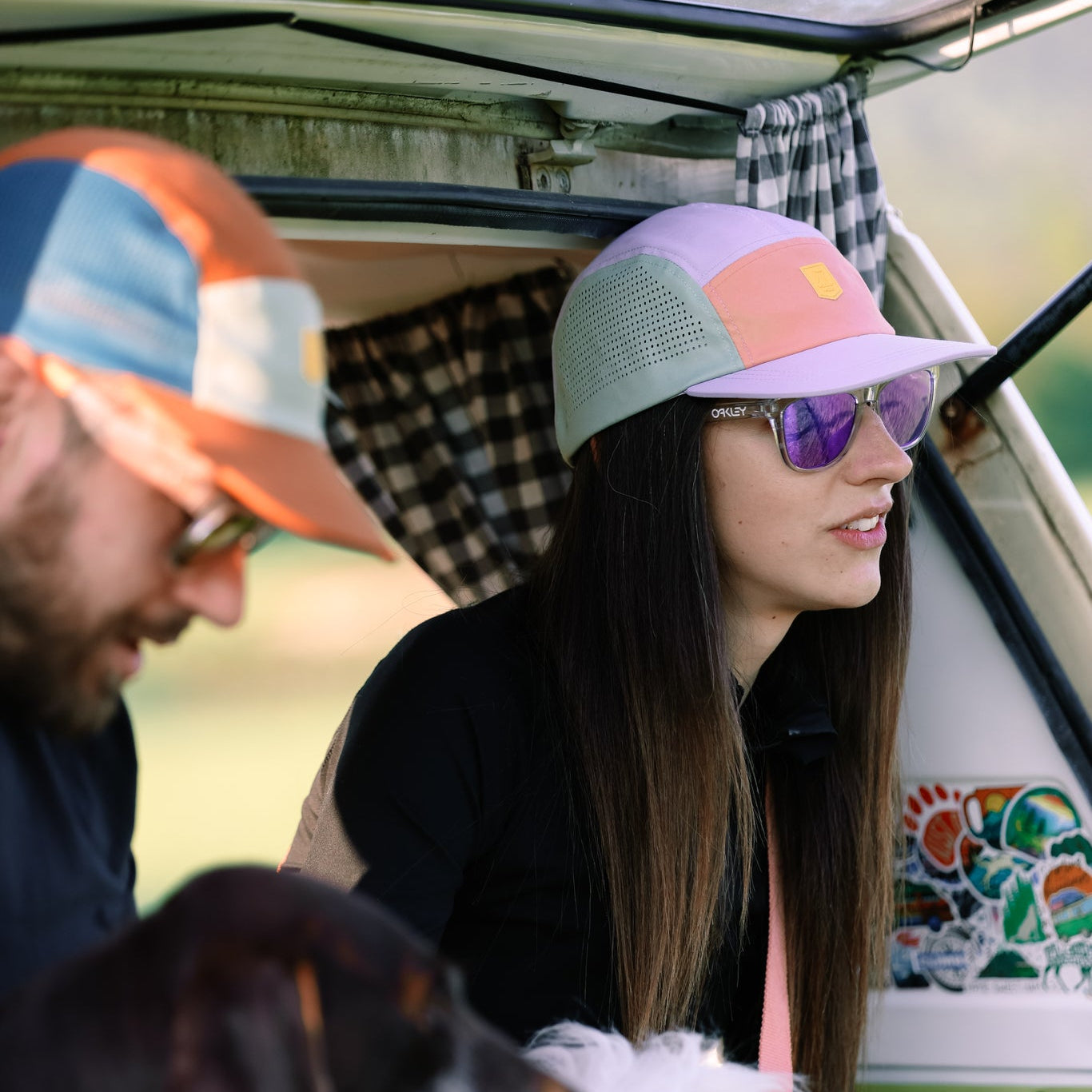 Two people relax in an open van with two dogs. The woman, in reflective sunglasses and an Alpha 360 Tech Cap Lavender, looks ahead as the man holds a dog. Their colorful hats and setting suggest a laid-back outdoor adventure.
