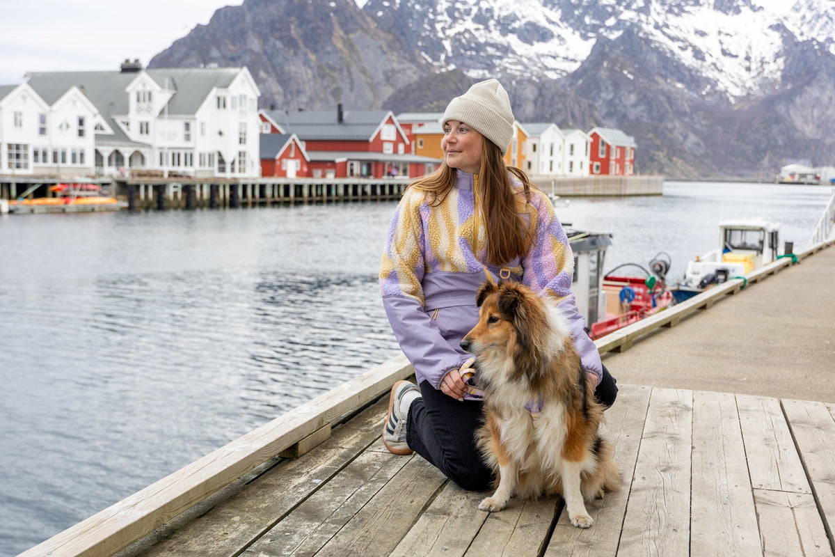 A woman and a Sheltie dog sit together on a wooden pier in front of a coastal village with snow-capped mountains.