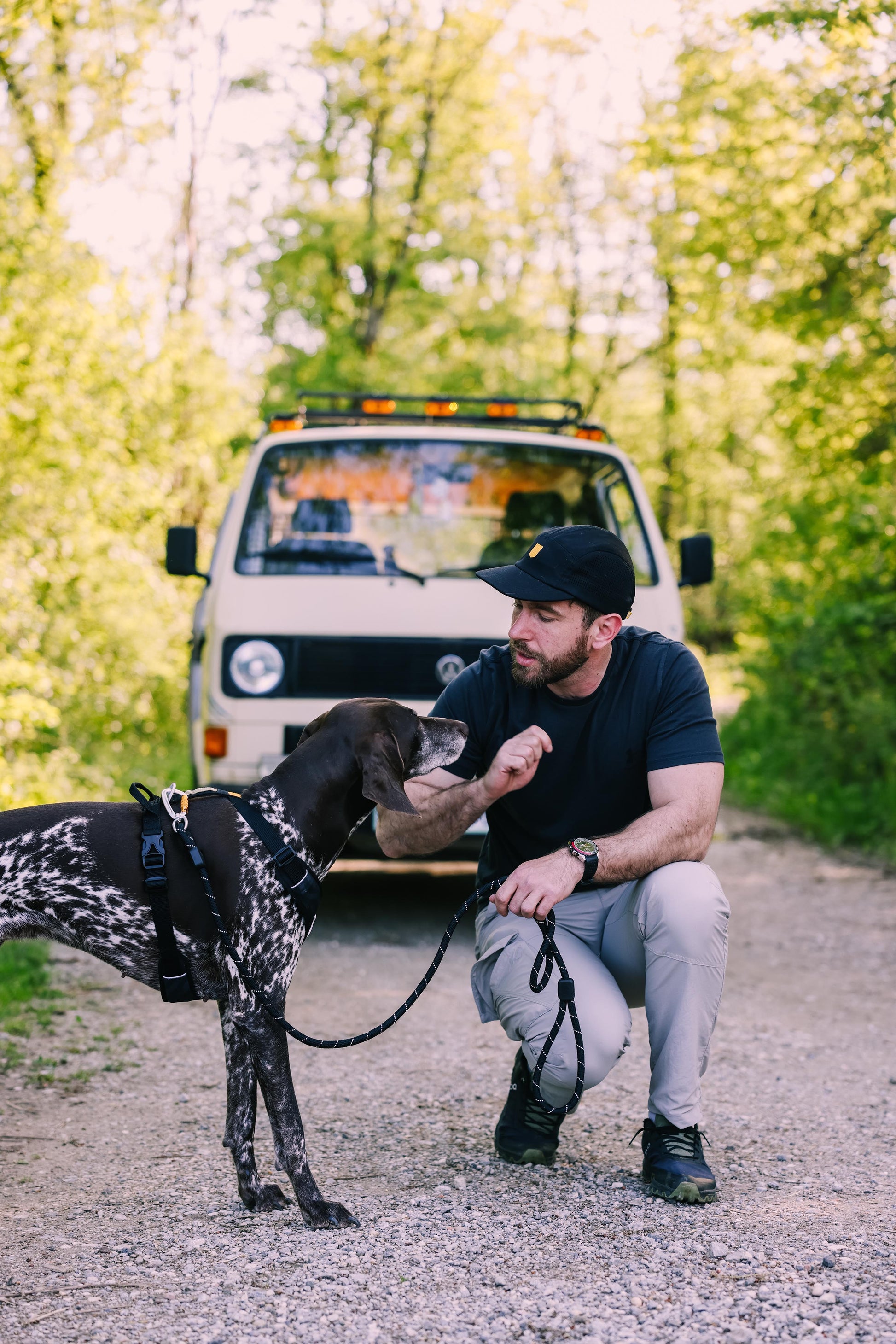 Man kneeling on a gravel road, petting a dog, with a beige van and trees in the background.