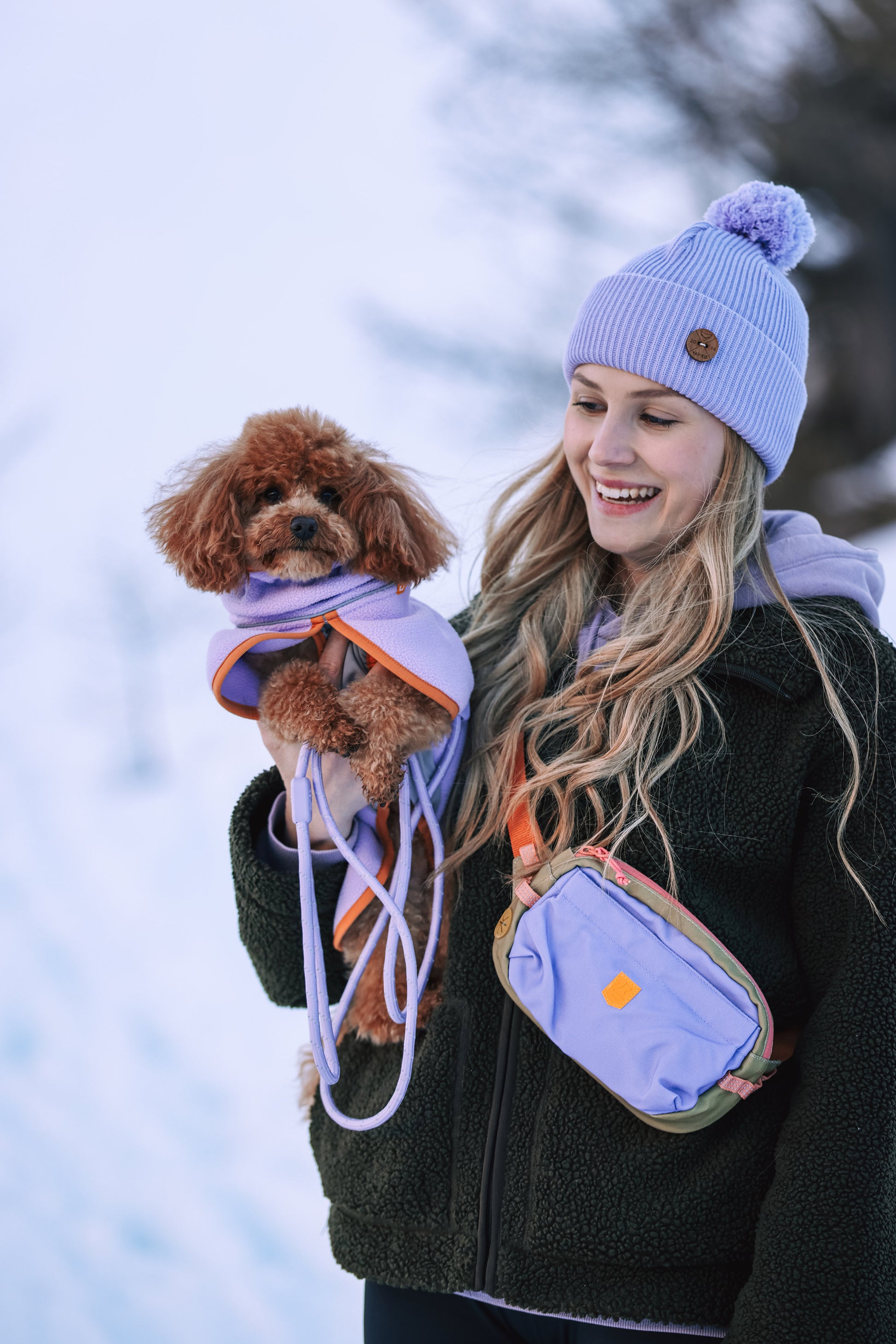 Smiling woman in a purple beanie holds a small brown dog dressed in a purple coat on a snowy day.