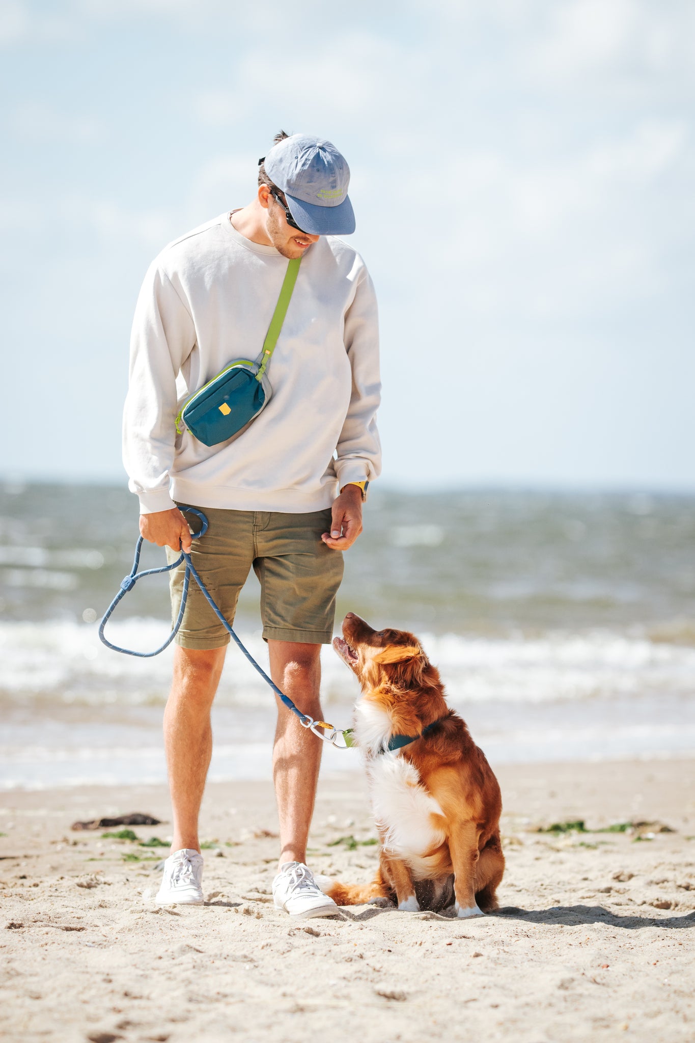 Man walking a brown dog on a leash at the beach, both looking at each other, with the ocean in the background.