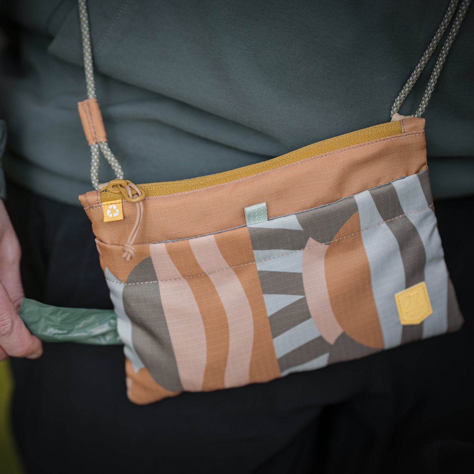 A person pulls a green bag from a small crossbody pouch with a geometric pattern.