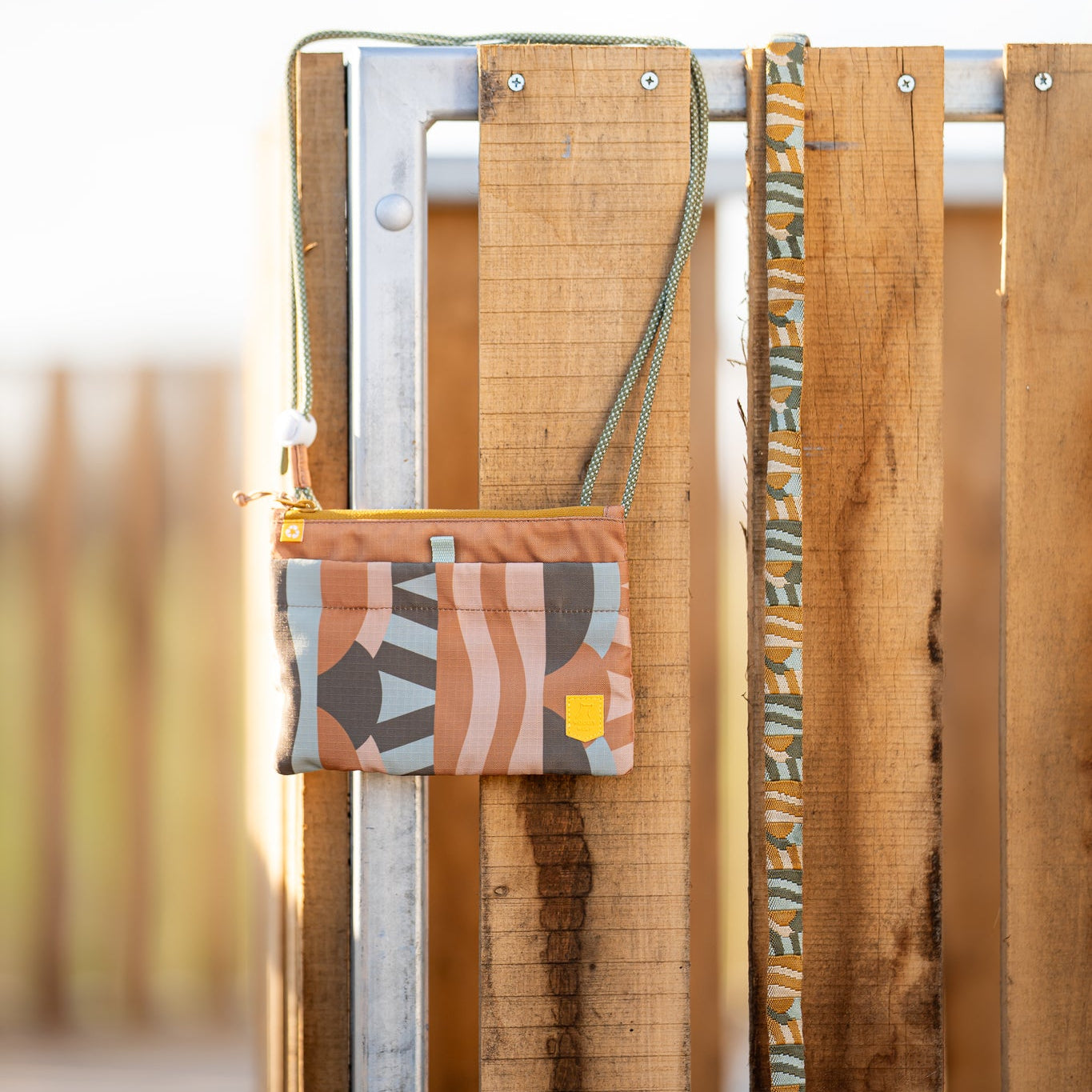 A small, patterned pouch with a strap hangs on a wooden and metal fence in bright sunlight.