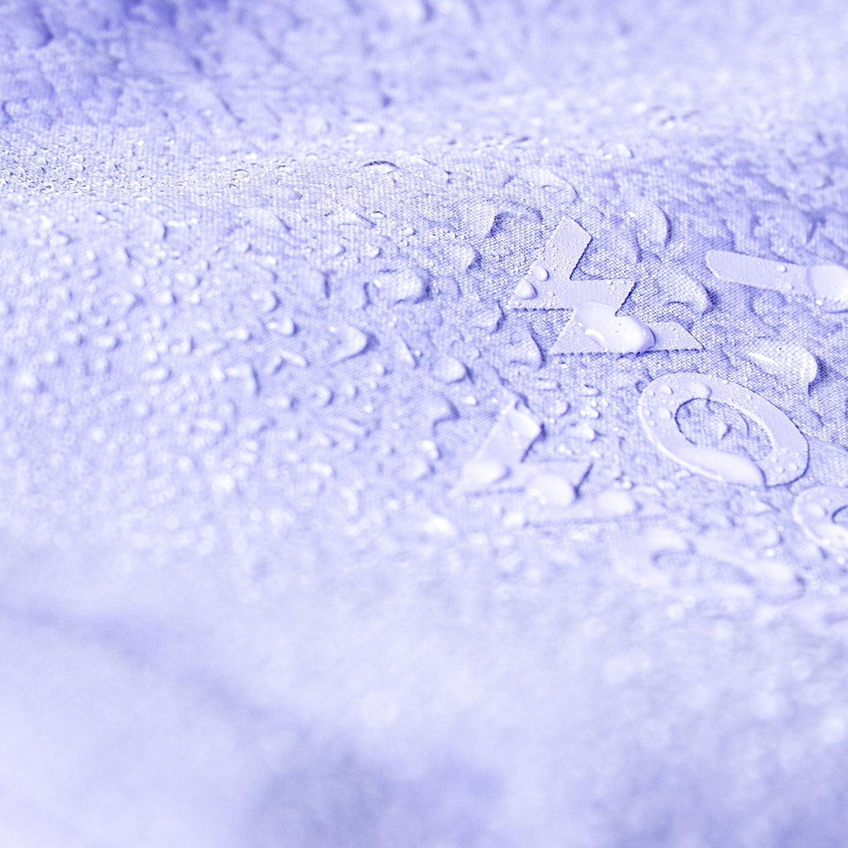 Close-up of water droplets on a textured, light purple surface with faintly visible letters underneath.