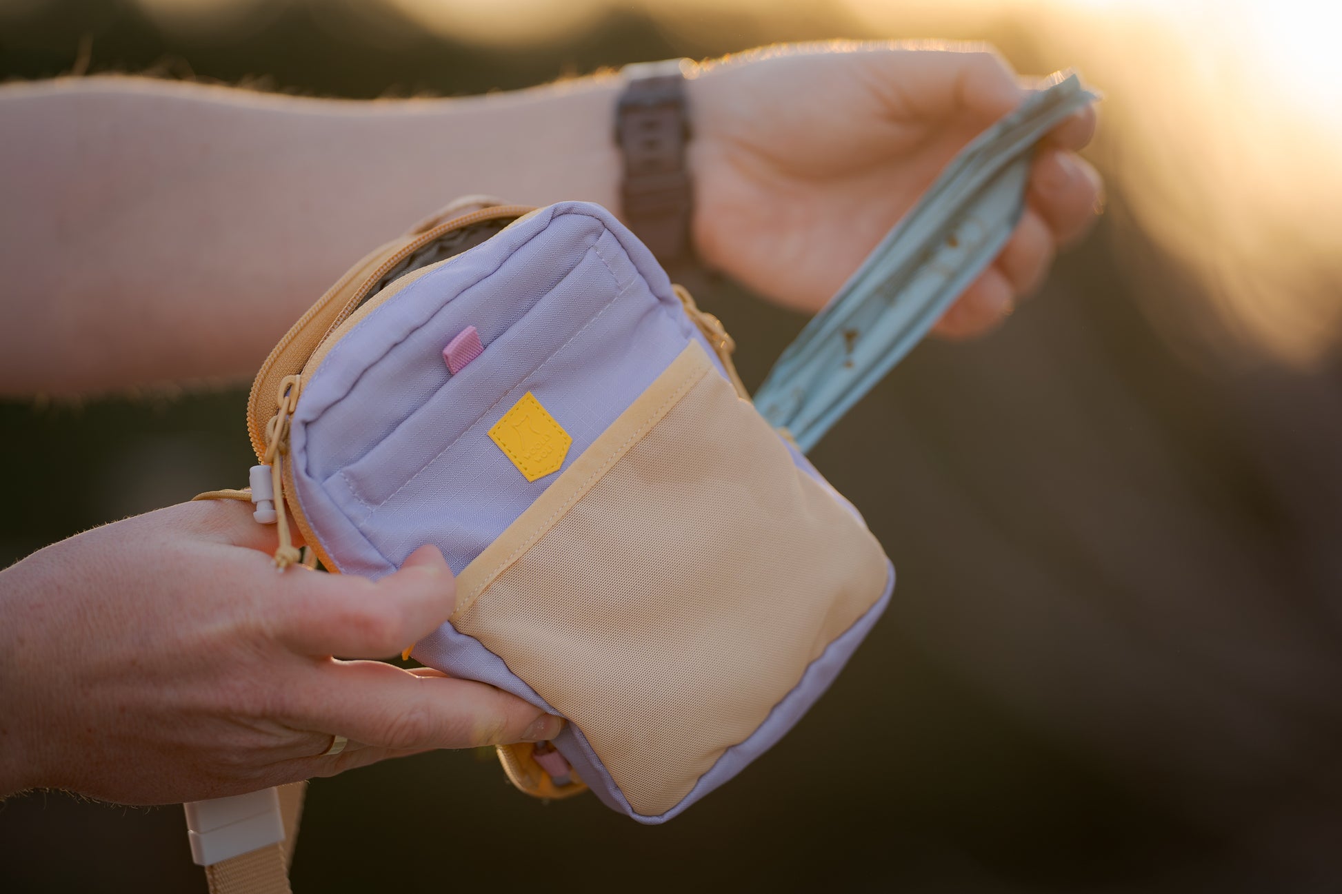 A person holds a small pastel crossbody bag and pulls out a teal face mask at sunset.