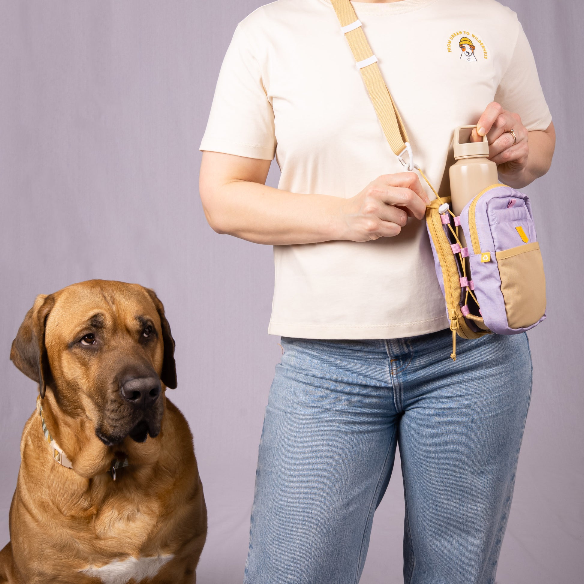 A person with a crossbody bag and water bottle stands next to a large brown dog against a plain background.