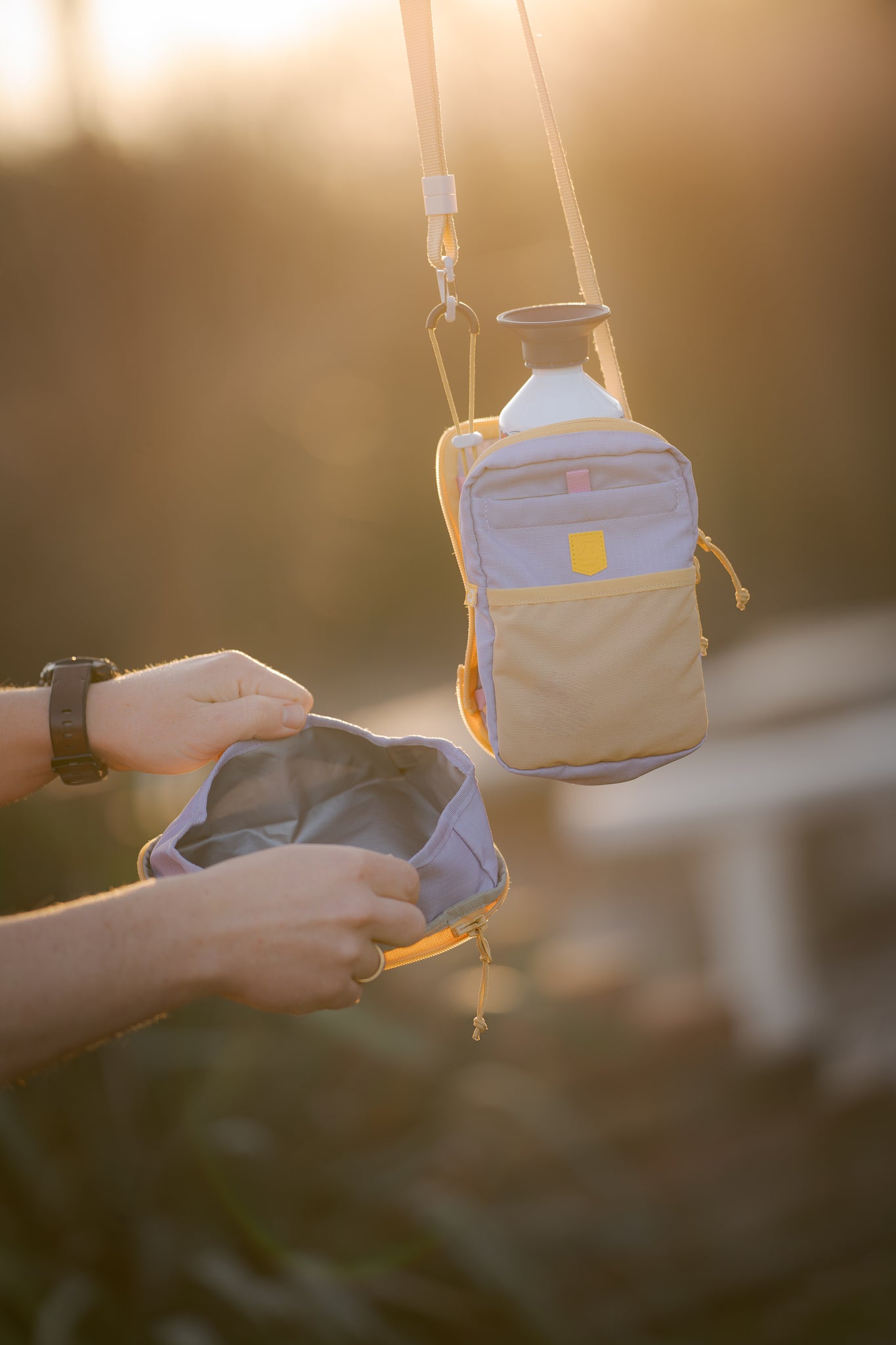 Two small bags, one hanging with a water bottle, another held open by a person, outdoors at sunset.
