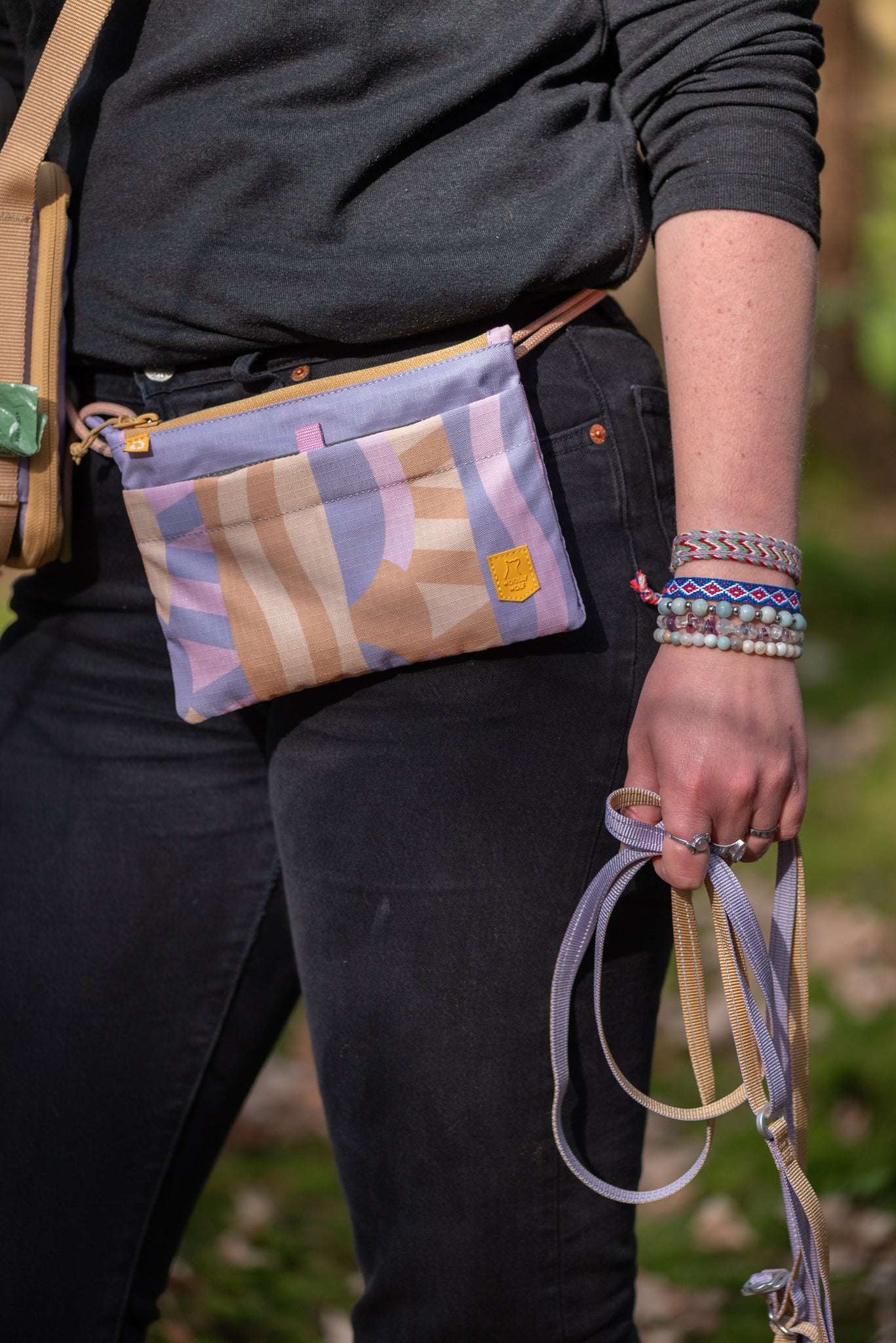 Person wearing black clothes with a pastel patterned fanny pack and holding woven leashes and beaded bracelets.