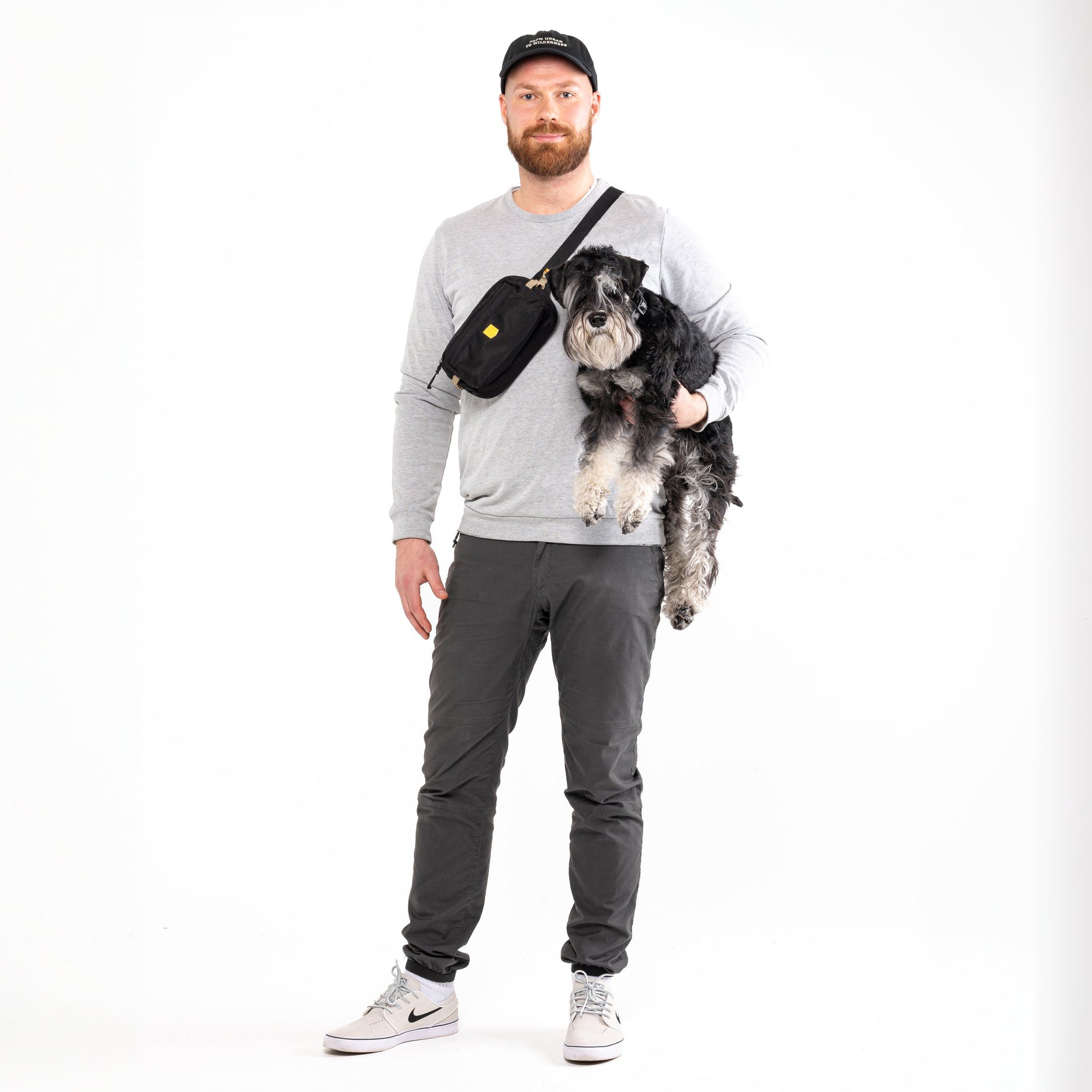 A man in a grey sweatshirt and black cap holds a small black dog and a black crossbody bag.