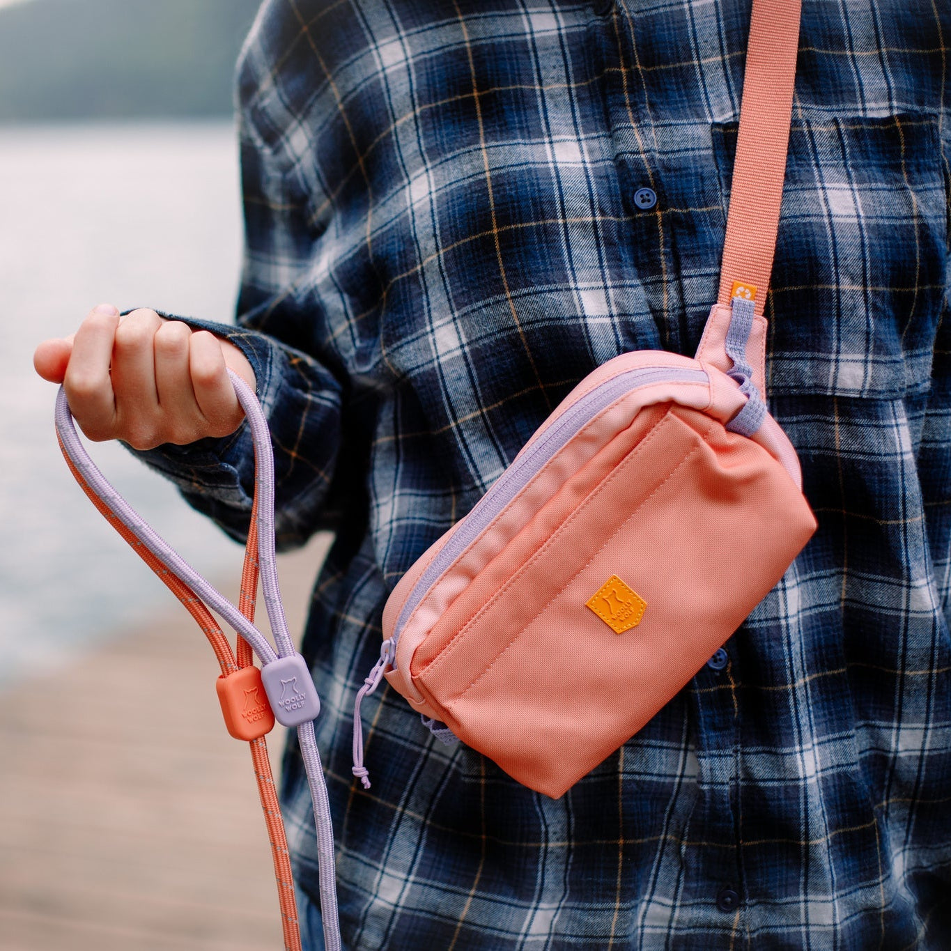 A person in a blue plaid shirt stands on a dock by the water, holding a pink leash and wearing the Alpha 360 Hip Pack in Salmon Pink. The background is slightly blurred, showing water and greenery in the distance.
