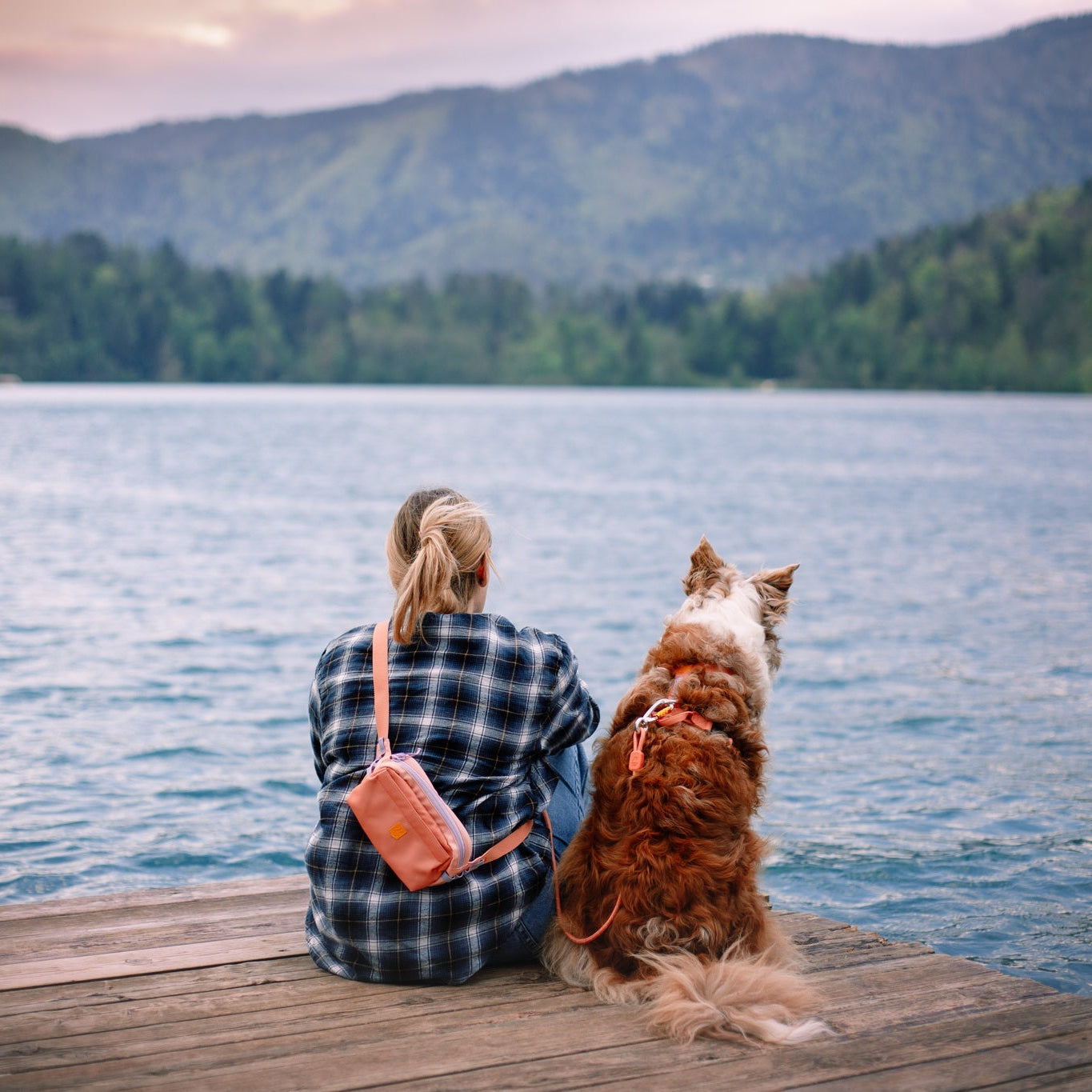 A person with an Alpha 360 Hip Pack in Salmon Pink sits on a wooden dock beside a brown and white dog, both gazing at a calm lake surrounded by forested hills and distant mountains.