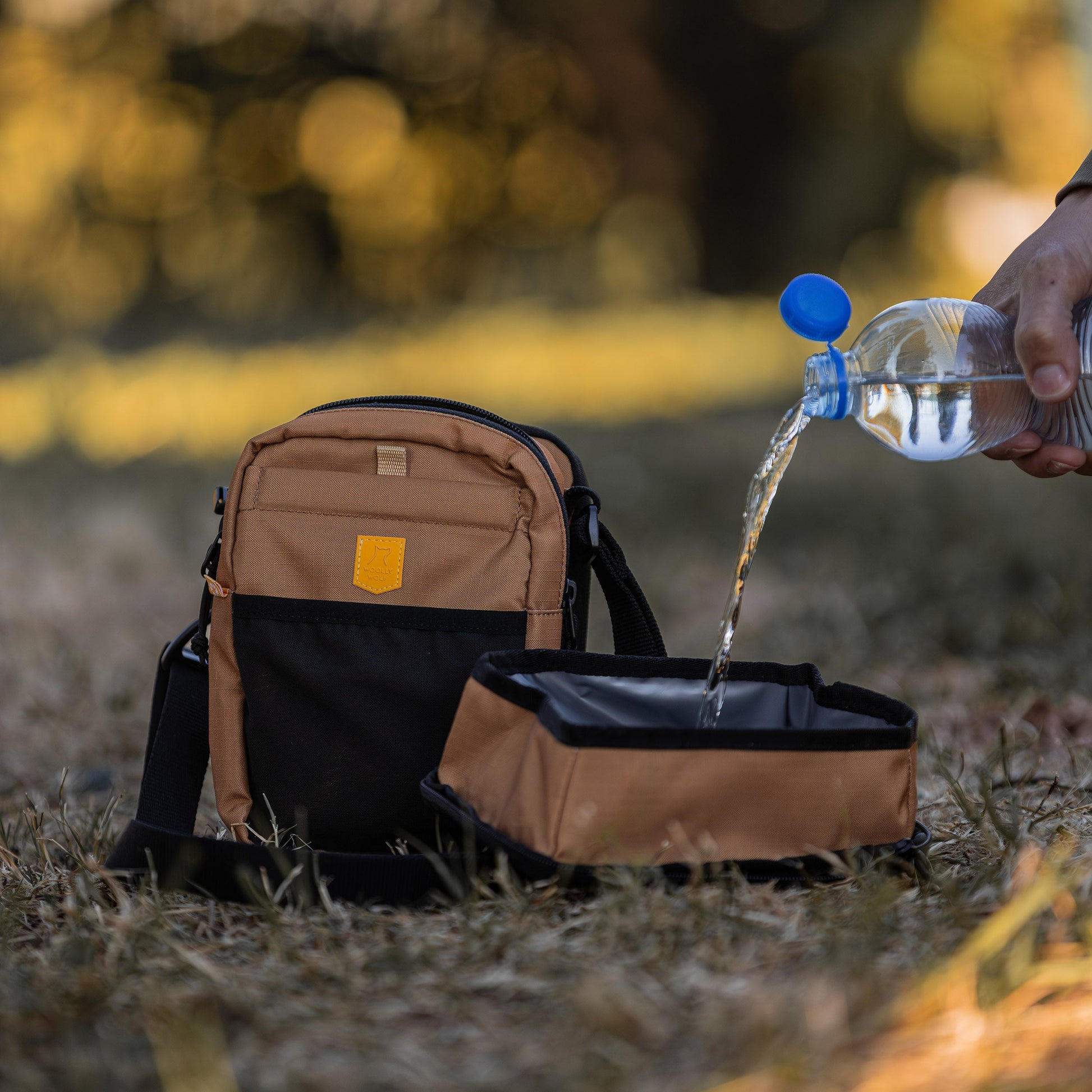 A hand pours water from a bottle into a portable brown and black pet bowl on grass beside a matching bag.