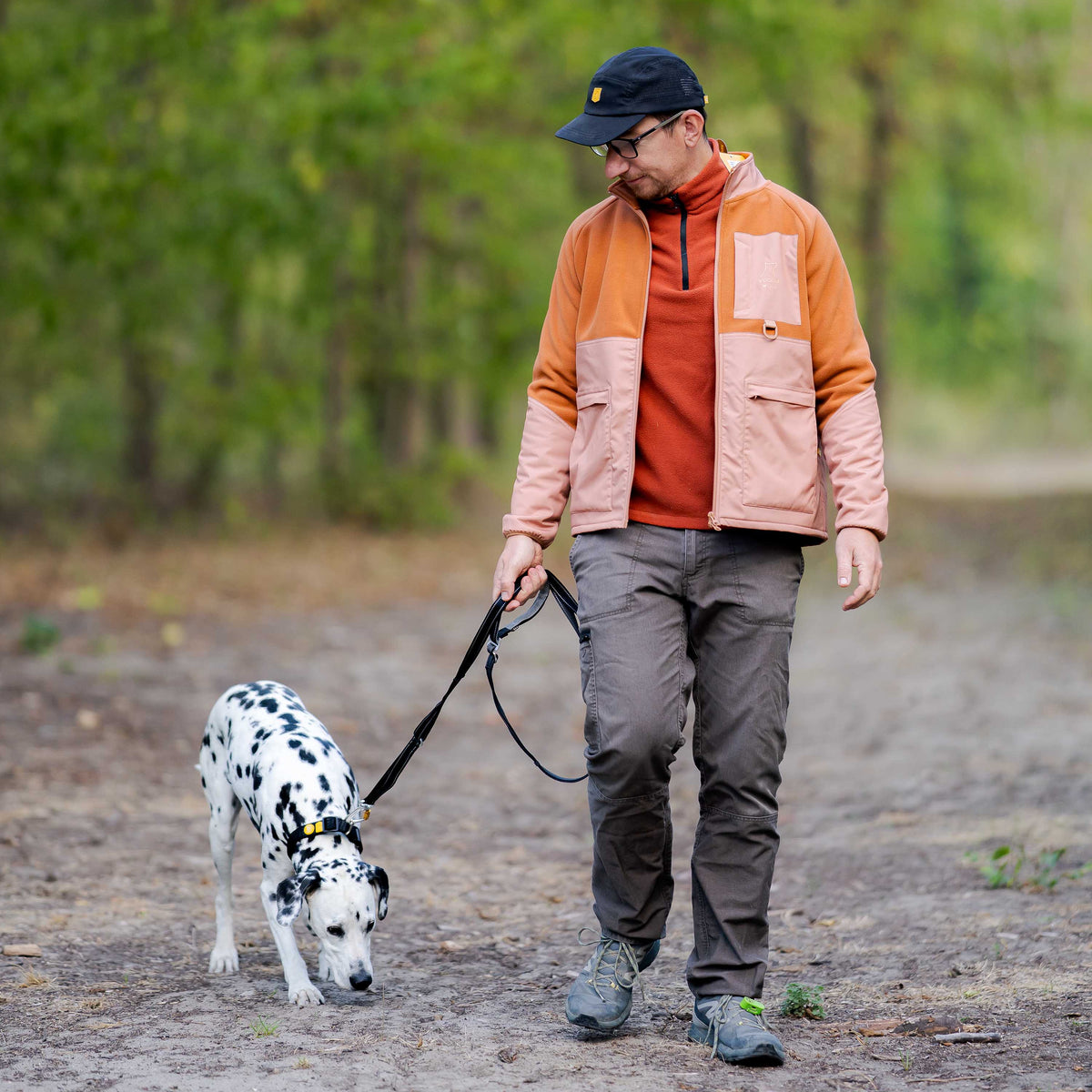 A man in a jacket and cap walks a Dalmatian on a dirt path in the woods.