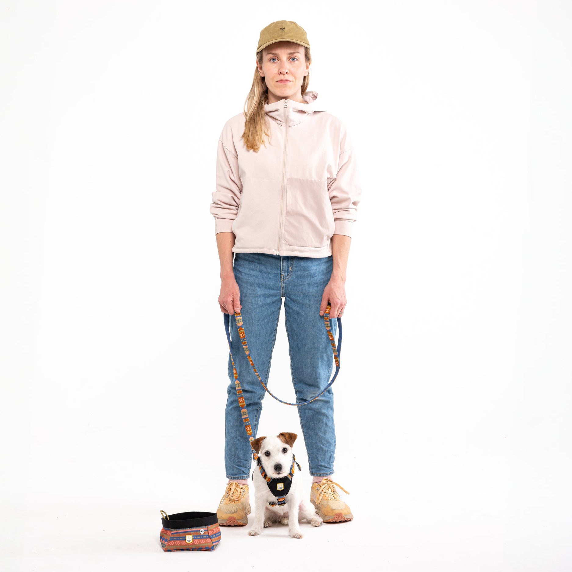 Woman in cap and jeans holds leash attached to dog. Collapsible bowl nearby.